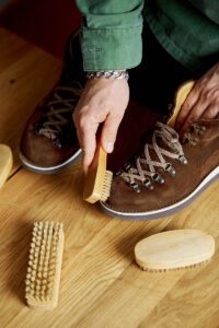 A person’s hands cleaning suede boots with a brush on a wooden background.